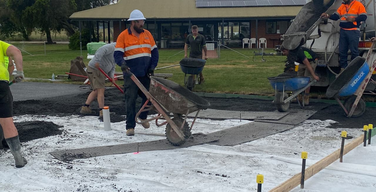 Crew wheelbarrowing permeable concrete from a mixer truck over rubber mats onto a fabric-lined base outside a single-storey building with solar panels