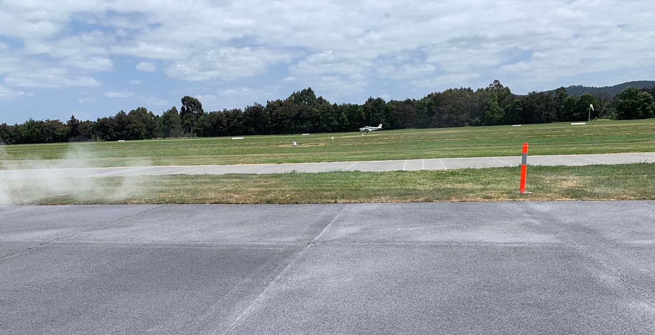 Light aircraft on the runway beyond a grey permeable concrete apron, with an orange marker post and a tree line behind