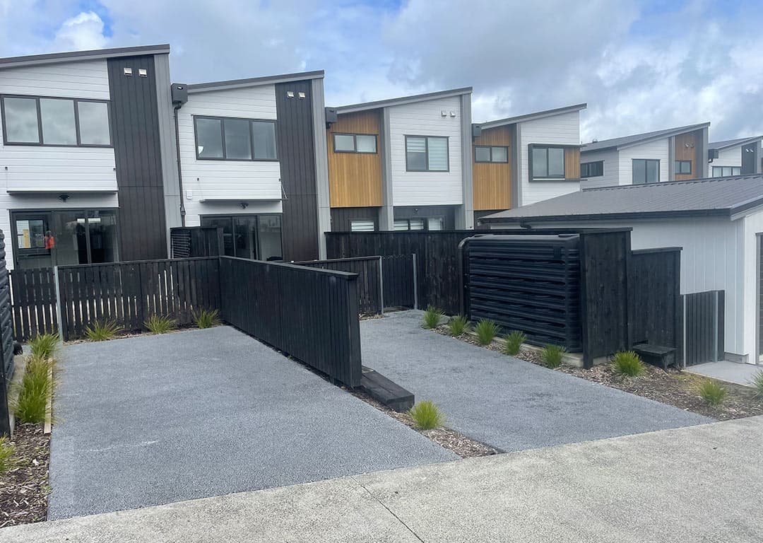 Two short grey permeable driveways side by side in front of new townhouses, divided by a black timber fence and edged with grasses