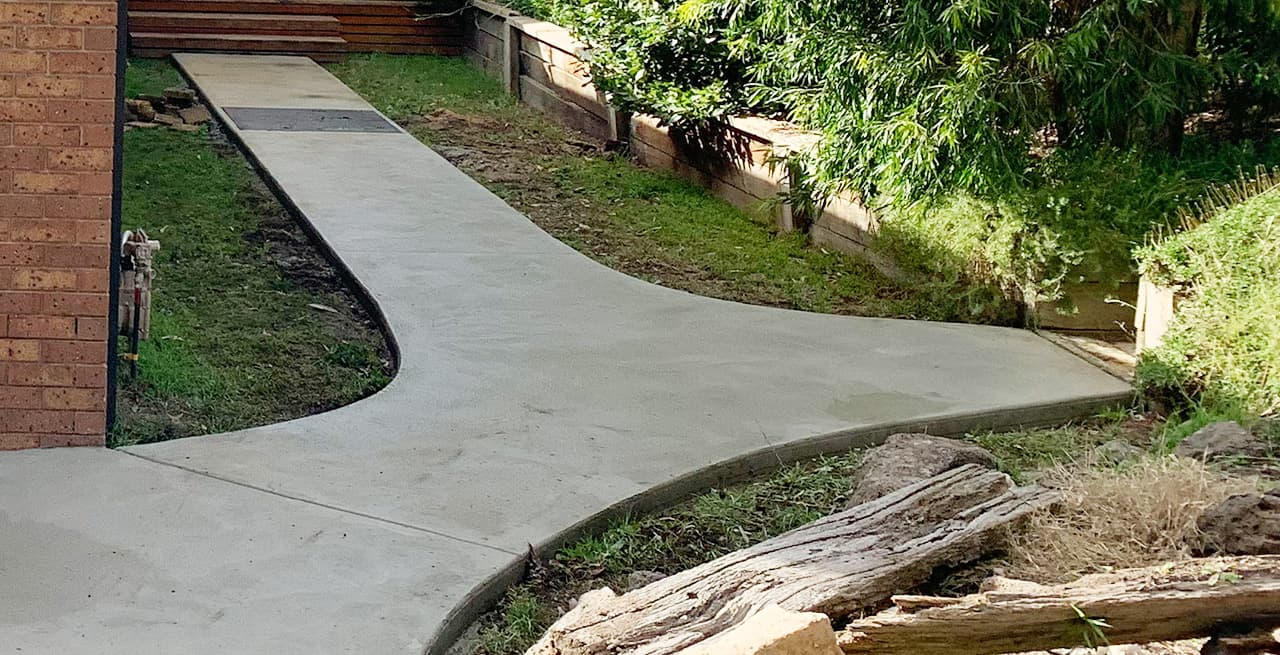 Pale grey poured path forking between a brick wall and a timber retaining wall, with lawn and a garden bed either side