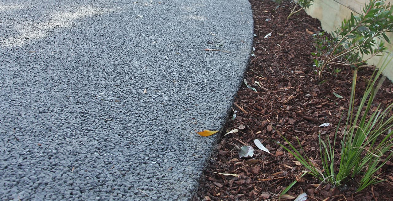 Close-up of the curved edge of a grey permeable concrete path meeting a mulched bed with young plants, a few fallen leaves on the surface
