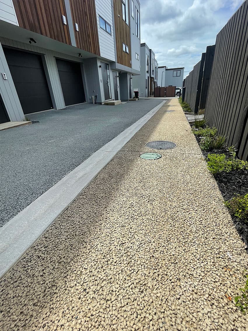 Grey shared driveway running past a row of townhouse garages, with a sandstone-toned open-textured path along the fence