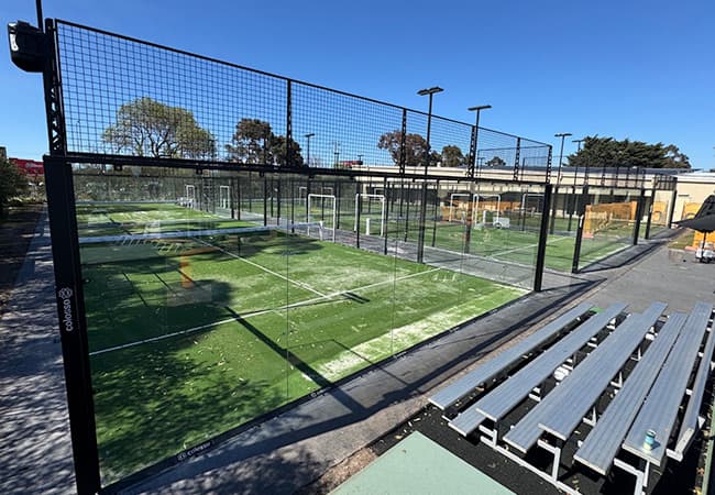 Padel courts behind glass and black steel mesh, with green synthetic turf and aluminium bench seating in front